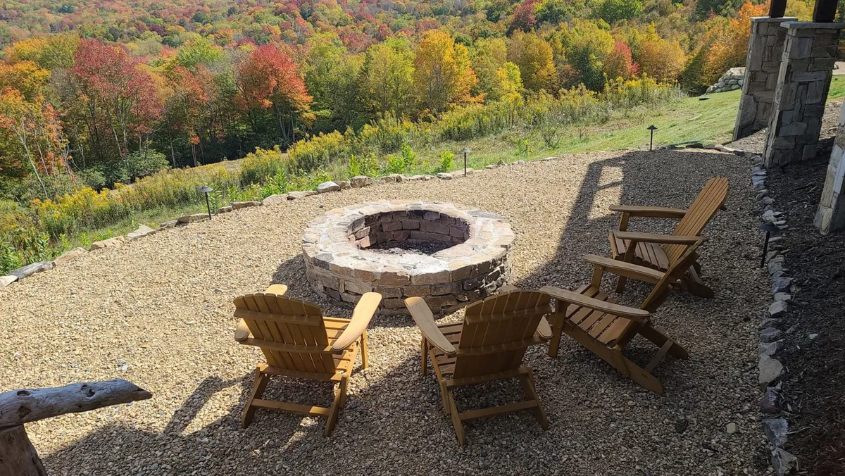 A cozy outdoor fire pit area surrounded by Adirondack chairs, set against a backdrop of vibrant fall foliage and expansive sunset views over the valley. The scene invites relaxation and gathering, with the fire pit as the central feature, perfect for storytelling and stargazing.