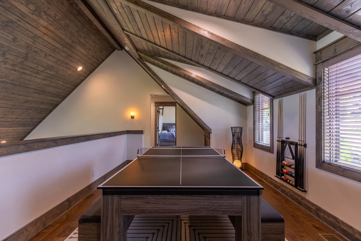 A spacious, light-filled recreation room featuring a ping pong table under a sloped wooden ceiling, with decorative lighting and billiard cues displayed on the wall. The playful atmosphere is enhanced by ample space for movement and socializing, while the overall setting is clean, inviting, and geared toward group activities.