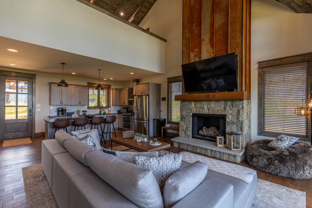 A luxurious open-concept interior featuring a stone fireplace, large sectional sofa, spacious kitchen island, and a dining area. The room is filled with natural light from large windows, which frame scenic views of the mountains beyond. The space is arranged for social gatherings, with cozy seating near the fireplace and the kitchen and dining area seamlessly connected.