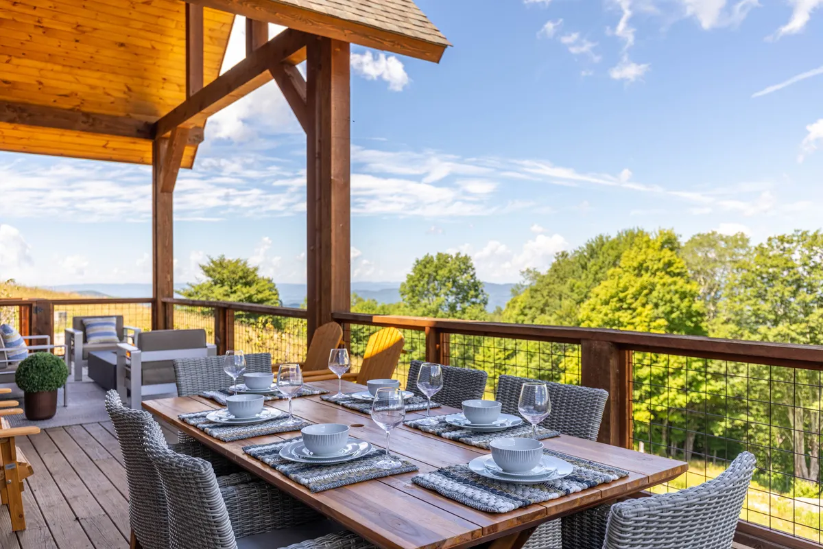 A wooden porch features a dining table set with dishes, framed by large mountain vistas in the background. The cheerful setup invites guests to enjoy meals together while soaking in panoramic views and fresh air.