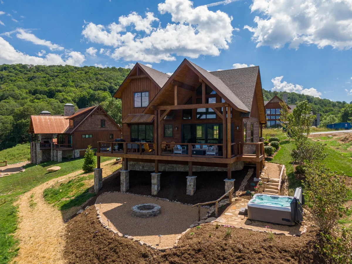 The image shows a spacious, newly built wooden cabin nestled among lush greenery on the ridgeline of Eagles Nest in Banner Elk, North Carolina. The exterior features a wrap-around porch perfect for relaxing, a bubbling hot tub with scenic mountain views, and a cozy fire pit area ideal for gathering under the stars. The overall mood is peaceful and inviting, with the natural beauty of the mountains complementing the home's charming design.